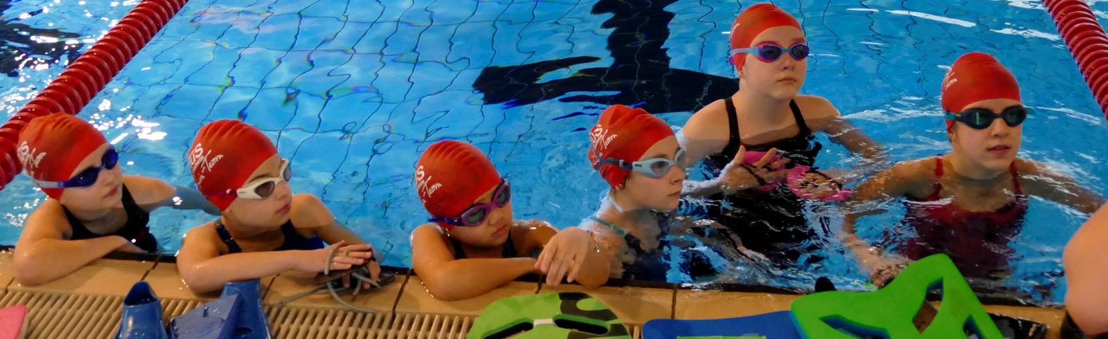 Sechs junge Schwimmerinnen mit roten Badekappen und Schwimmbrillen lehnen am Beckenrand eines Schwimmbeckens.