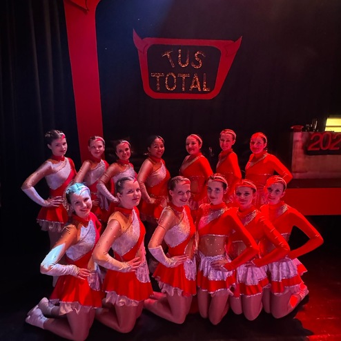 Gruppe von Tänzerinnen in roten Kostümen posiert auf der Bühne vor einem dekorierten Hintergrund mit „TuS TOTAL“.
