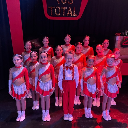 Gruppe von Kindern in roten Kostümen mit Glitzer, stehen vor schwarzem Hintergrund mit „TUS TOTAL“ Schriftzug.