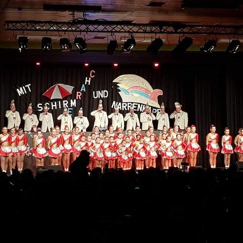 Bühne mit Gruppen von Tänzern in roten und weißen Kostümen sowie einem Ensemble in Anzügen, umgeben von Dekorationen.