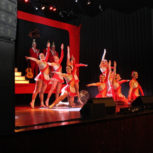 Tänzerinnen in roten Kostümen präsentieren eine choreografierte Darbietung auf einer Bühne mit Bühnenbild.