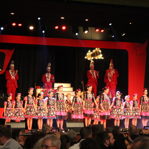 Bühne mit Kindern in bunten Kleidern, die Skelettmuster zeigen; im Hintergrund applaudierende Personen in roten Anzügen.