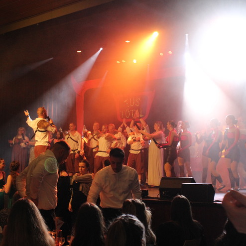 Bühne mit tanzenden Darstellern und Publikum, von bunten Lichtern und Nebel umgeben, bei einer lebhaften Aufführung.