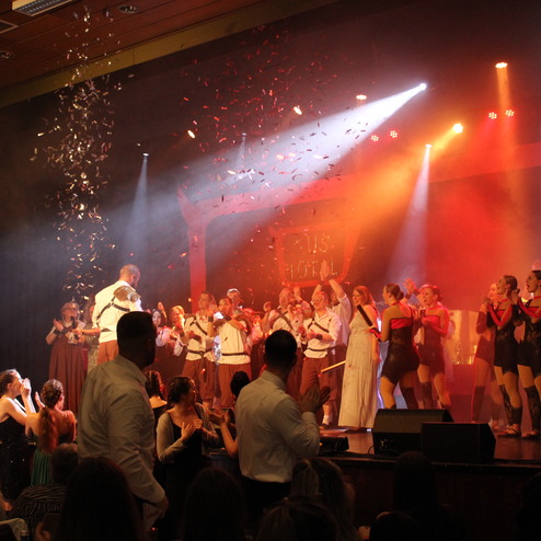 Bühnenaufführung mit tanzenden Darstellern, Konfetti und Applaus vor bunten Lichtern in einem festlichen Saal.