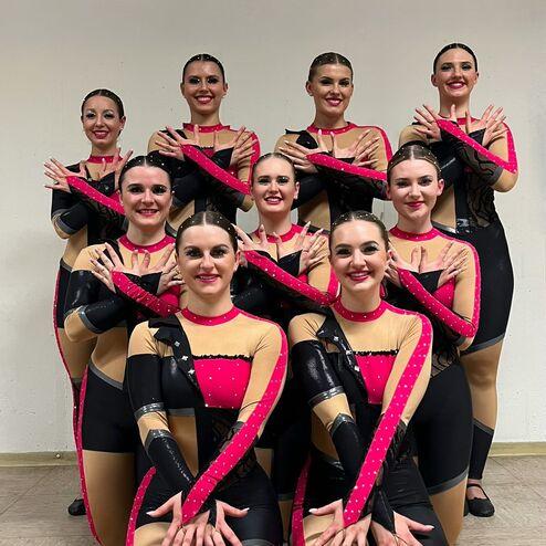 Neun Tänzerinnen in auffälligen, schwarz-pinken Kostümen posieren in mehreren Reihen für ein Gruppenfoto.