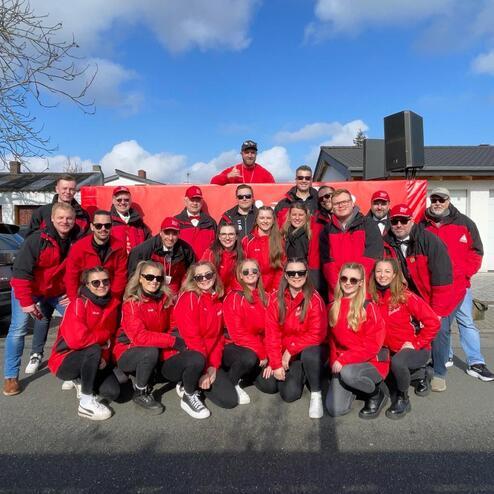 Gruppe von 25 Personen in roten Jacken posiert lächelnd vor blauer Himmel mit Wolken, einige tragen Sonnenbrillen.