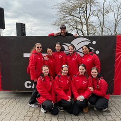 Gruppe von zehn Frauen in roten Jacken posiert vor einem schwarzen Banner, umgeben von B&auml;umen und parkenden Autos.