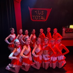Gruppe von T&auml;nzerinnen in roten Kost&uuml;men posiert auf der B&uuml;hne vor einem dekorierten Hintergrund mit &bdquo;TuS TOTAL&ldquo;.