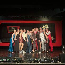 Gruppe von 16 Personen in festlicher Kleidung l&auml;chelt auf einer B&uuml;hne mit schwarz-rotem Hintergrund.