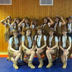 Gruppe von T&auml;nzerinnen in Cowboy-Outfits mit H&uuml;ten, posierend in einem Holzraum mit blauer Matte und rotem Rand.