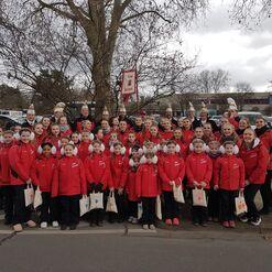 Gruppe von T&auml;nzern in roten Jacken mit Beuteln, versammelt unter einem Baum, vor einer Veranstaltung.
