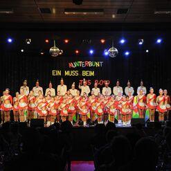 Auftritt einer Tanzgruppe in bunten Kost&uuml;men auf einer B&uuml;hne mit festlichem Dekor und Publikum im Vordergrund.