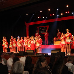 Gruppe von T&auml;nzern in roten Kost&uuml;men auf einer B&uuml;hne, Zuschauer sitzen an Tischen im Vordergrund.