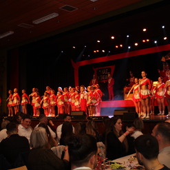 Tanzende Kost&uuml;me in Rot und Silber auf der B&uuml;hne, Publikum sitzt an Tischen und beobachtet die Auff&uuml;hrung.