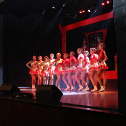 Tanzgruppe in roten Kost&uuml;men steht auf einer B&uuml;hnenb&uuml;hne vor einem schwarzen Vorhang mit B&uuml;hnenbeleuchtung.