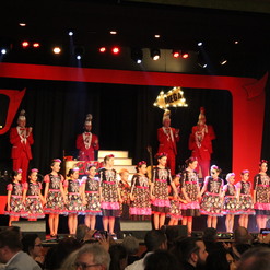 B&uuml;hne mit Kindern in bunten Kleidern, die Skelettmuster zeigen; im Hintergrund applaudierende Personen in roten Anz&uuml;gen.