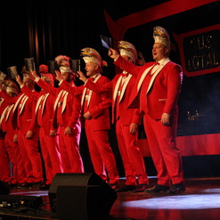 Gruppe von M&auml;nnern in roten Anz&uuml;gen mit gl&auml;nzenden H&uuml;ten, die auf einer B&uuml;hne stehen und bl&auml;tternde Programme halten.