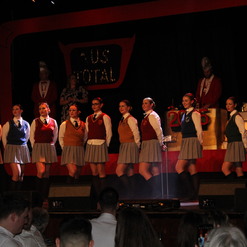 Gruppenauftritt von neun T&auml;nzerinnen in Schuluniformen auf einer B&uuml;hne mit dekortivem Hintergrund.