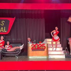 Auf der B&uuml;hne posieren T&auml;nzerinnen in rot-silbernen Kost&uuml;men vor einer dekorierten Kulisse mit Schriftzug &bdquo;TUS TOTAL&ldquo;.