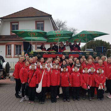 Gruppe von Mädchen in roten Jacken posiert vor einem Wagen mit Sonnenschirmen und einem Gebäude im Hintergrund.