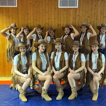 Gruppe von T&auml;nzerinnen in Cowboy-Outfits mit H&uuml;ten, posierend in einem Holzraum mit blauer Matte und rotem Rand.