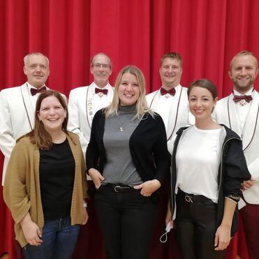 Gruppe von sechs Personen in Abendgarderobe, lächelnd vor rotem Vorhang. Drei Männer tragen weiße Sakkos, drei Frauen farbige Kleidung.