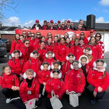 Gruppe von Mädchen und Erwachsenen in roten Jacken und Mützen posiert gemeinsam vor einem Hintergrund mit blauem Himmel.