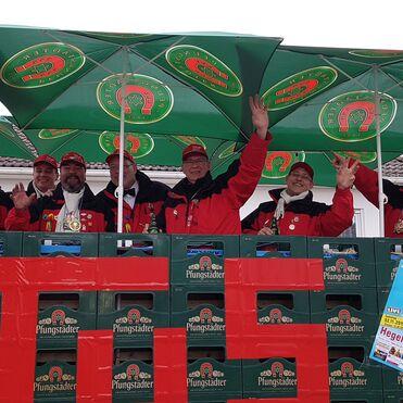 Sechs Personen in roten Jacken winken hinter einem Kastenstapel mit einem Pfungstädter-Logo und Sonnenschirmen.