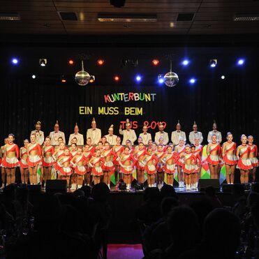 Auftritt einer Tanzgruppe in bunten Kostümen auf einer Bühne mit festlichem Dekor und Publikum im Vordergrund.