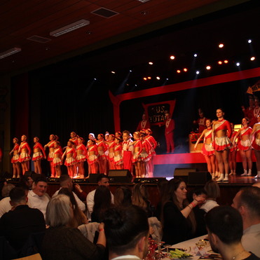 Tanzende Kost&uuml;me in Rot und Silber auf der B&uuml;hne, Publikum sitzt an Tischen und beobachtet die Auff&uuml;hrung.