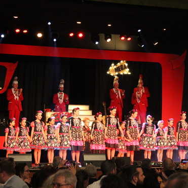 B&uuml;hne mit Kindern in bunten Kleidern, die Skelettmuster zeigen; im Hintergrund applaudierende Personen in roten Anz&uuml;gen.