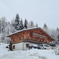 Holzhütte mit Balkon, umgeben von schneebedeckten Bäumen und einem verschneiten Parkplatz im Vordergrund.