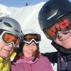 Drei Skifahrer mit Helmen und Brillen lächeln vor verschneiter Bergkulisse in einem sonnigen Skigebiet.