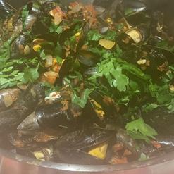 Edelstahlpfanne mit Muscheln, Tomaten und frischer Petersilie auf einem Tisch. Im Hintergrund ist ein Handansatz sichtbar.