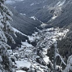 Schneebedeckte Berge umgeben ein Dorf im Tal, mit verschneiten Häusern und Tannenbäumen in der Landschaft.