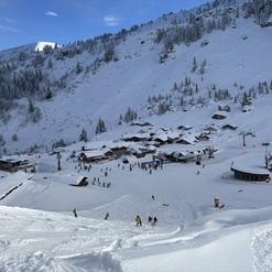 Panorama eines schneebedeckten Skigebiets mit Skifahrern und Hütten unter blauem Himmel.