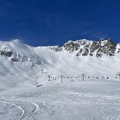 Schneebedeckte Berge und Skilifte unter blauem Himmel, mit Skifahrern auf der Piste im Vordergrund.