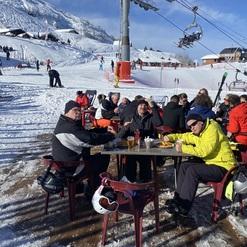 Skifahrer sitzen in einer Sonnenterrasse mit Getränken und Essen, umgeben von Schnee und Skifahrern im Hintergrund.