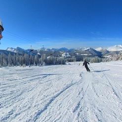 Skifahrer fährt eine verschneite Piste mit Bergpanorama und blauen Himmel im Hintergrund.