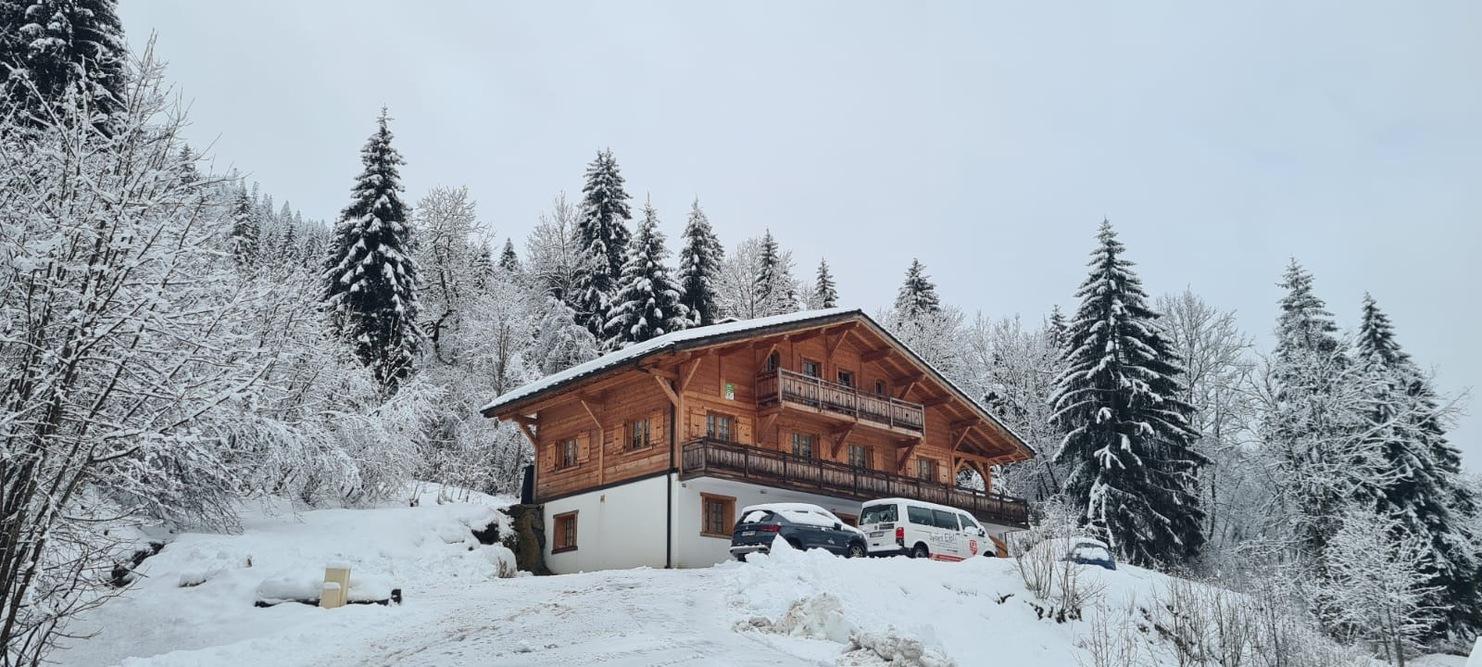 Holzhütte mit Balkon, umgeben von schneebedeckten Bäumen und einem verschneiten Parkplatz im Vordergrund.