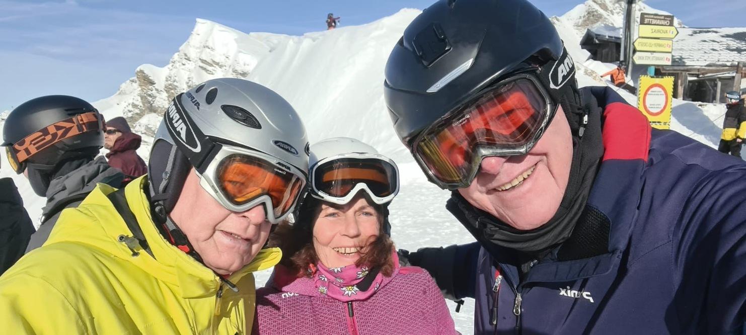 Drei Skifahrer mit Helmen und Brillen lächeln vor verschneiter Bergkulisse in einem sonnigen Skigebiet.