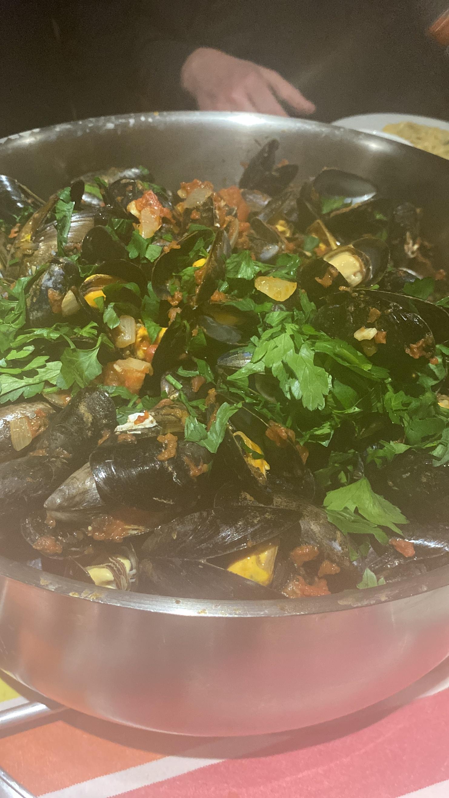 Edelstahlpfanne mit Muscheln, Tomaten und frischer Petersilie auf einem Tisch. Im Hintergrund ist ein Handansatz sichtbar.