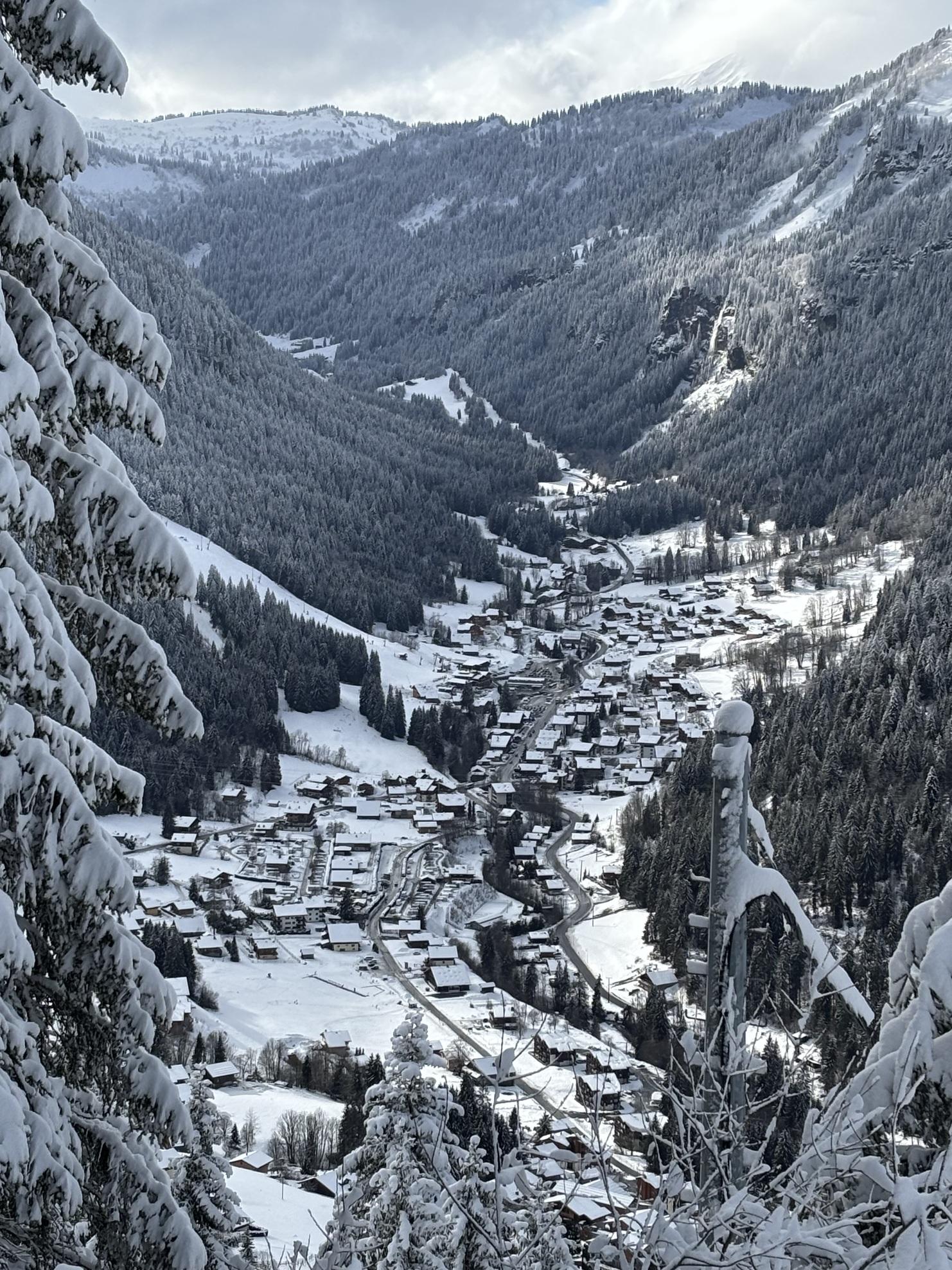 Schneebedeckte Berge umgeben ein Dorf im Tal, mit verschneiten Häusern und Tannenbäumen in der Landschaft.