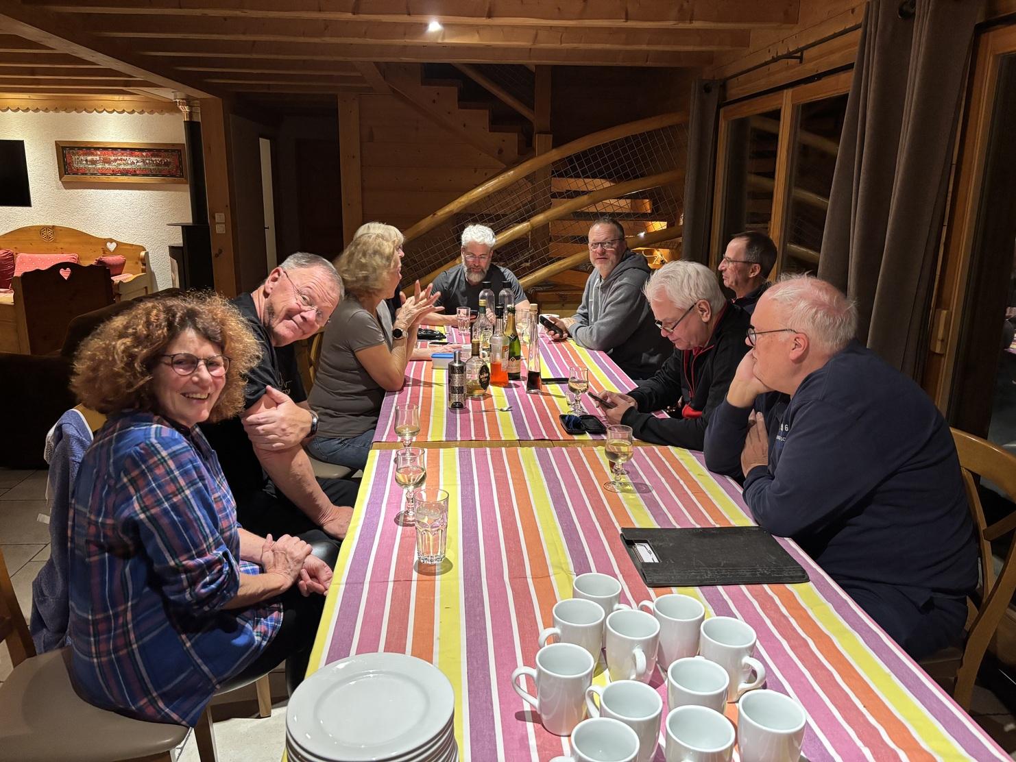 Geselliges Beisammensein am Tisch mit verschiedenen Personen, Getränken und Geschirr in einer holzverkleideten Umgebung.