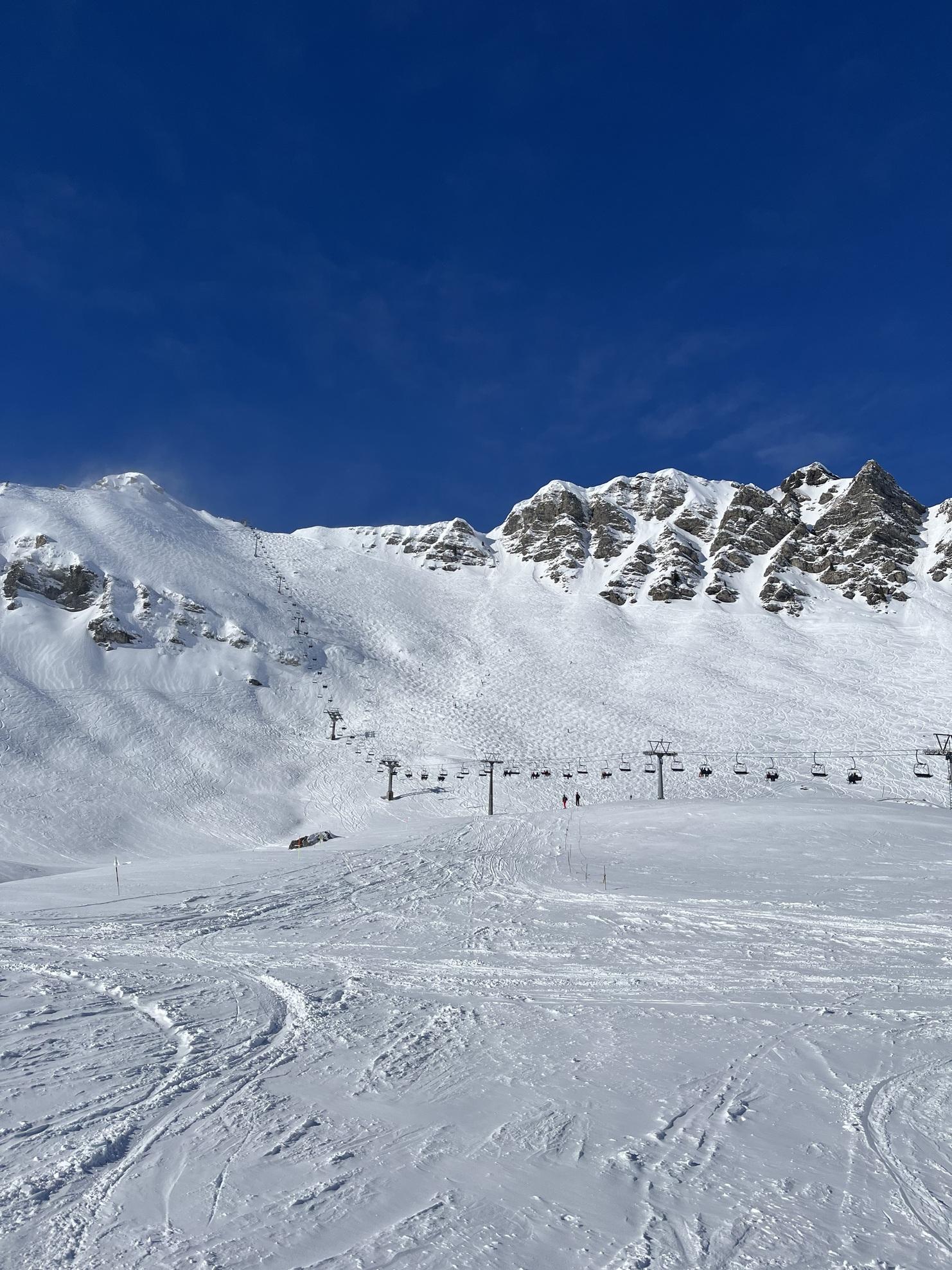 Schneebedeckte Berge und Skilifte unter blauem Himmel, mit Skifahrern auf der Piste im Vordergrund.