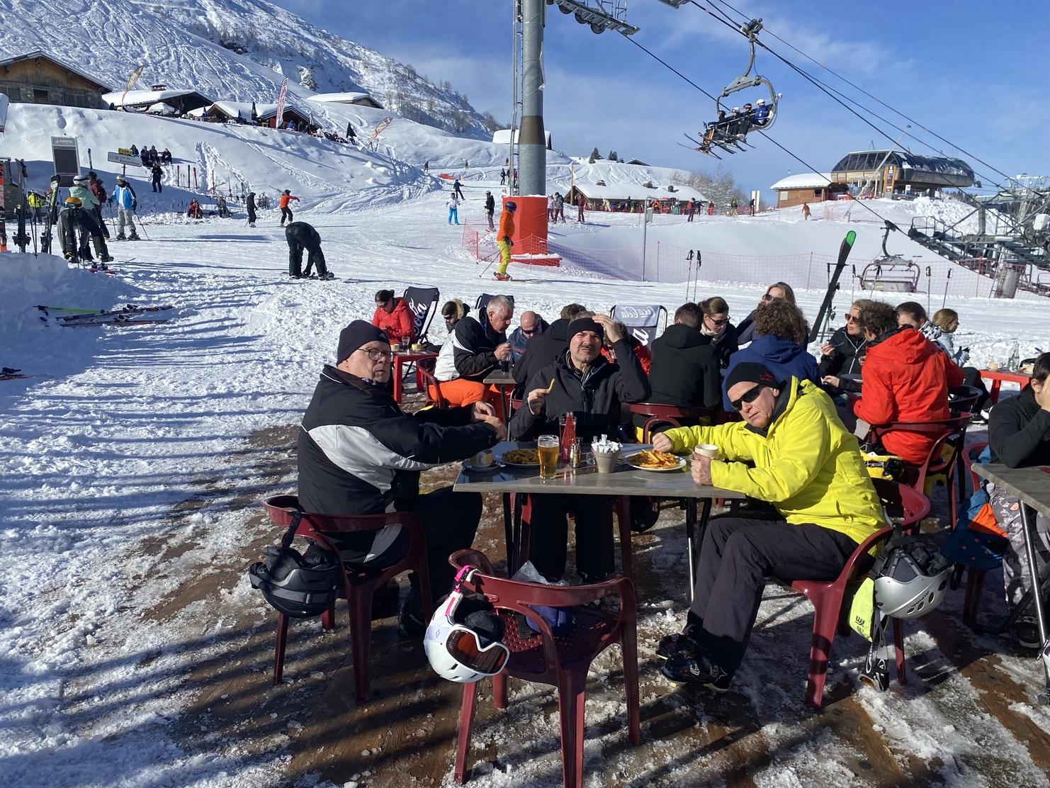 Skifahrer sitzen in einer Sonnenterrasse mit Getränken und Essen, umgeben von Schnee und Skifahrern im Hintergrund.