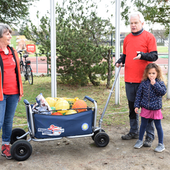 Zwei Erwachsene und ein Kind stehen an einem Sportplatz neben einem Wagen mit verschiedenen Sportbällen.