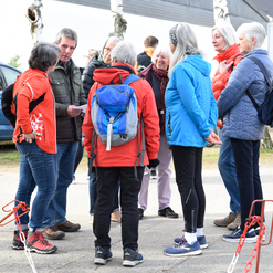Gruppe von acht Menschen in sportlicher Kleidung steht im Gespräch auf einem Weg, umgeben von Bäumen und Autos.