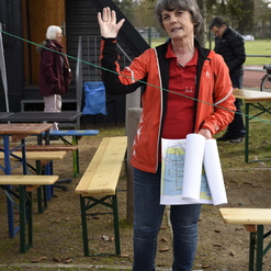 Frau mit Karten in der Hand, die im Freien spricht, während im Hintergrund Tische und andere Personen zu sehen sind.