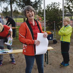 Lachende Frau in roter Jacke hält ein Blatt Papier, während ein Kind in grüner Jacke daneben steht. Andere Personen im Hintergrund.